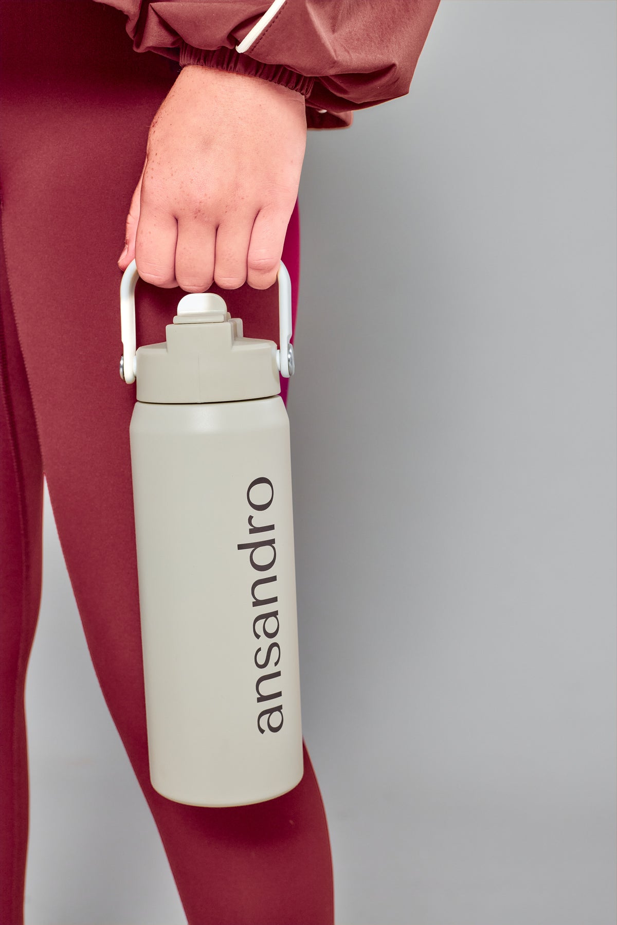 A person in burgundy activewear holds a light gray insulated water bottle with a white handle and the brand name 'ansandro' printed vertically in black on a plain gray background.