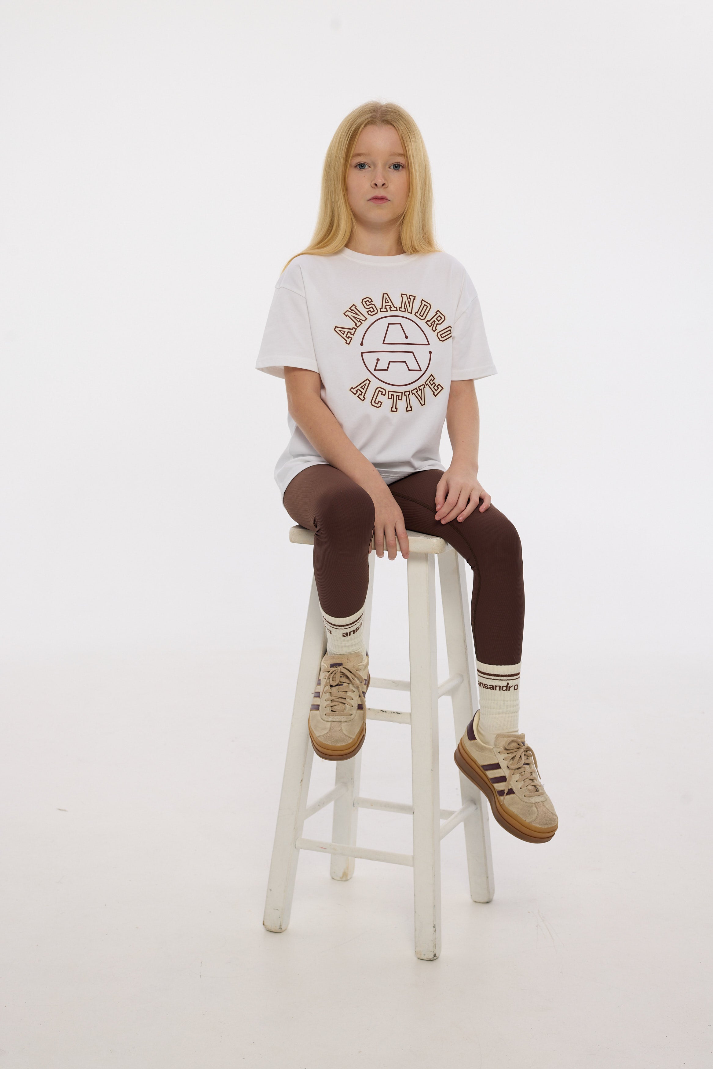 Person sitting on a stool wearing a white t-shirt with text and graphics, brown pants, and beige shoes.