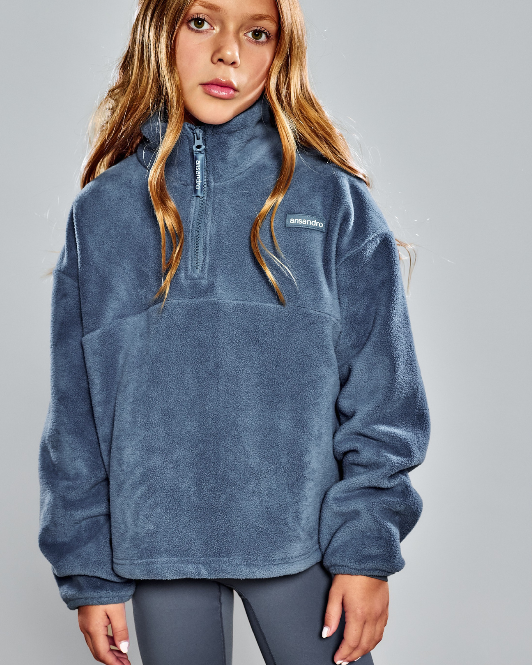 A young girl wearing a slate blue fleece half-zip pullover sweatshirt with a small white Patagonia logo on the chest, paired with gray leggings, against a solid gray studio background.