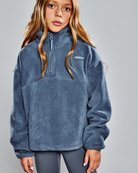 A young girl wearing a slate blue fleece half-zip pullover sweatshirt with a small white Patagonia logo on the chest, paired with gray leggings, against a solid gray studio background.