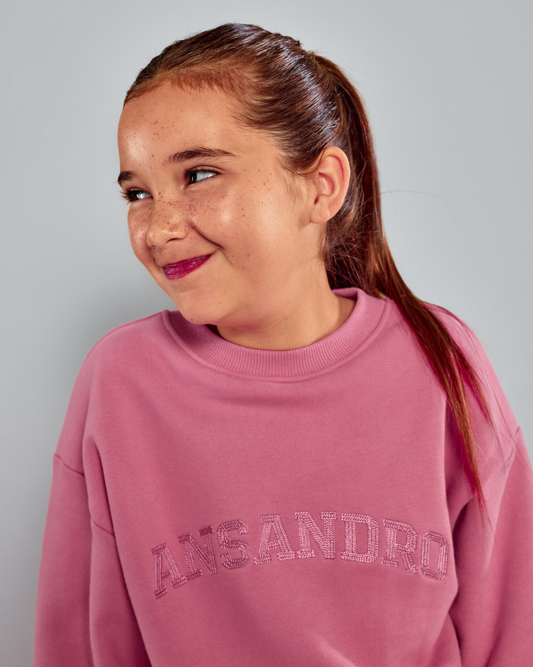 A young girl with her hair in a ponytail wearing a dusty pink crewneck sweatshirt with 'ANSANDRO' printed across the chest in a matching pink sequin or glittery font, set against a solid light gray background.