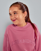 A young girl with her hair in a ponytail wearing a dusty pink crewneck sweatshirt with 'ANSANDRO' printed across the chest in a matching pink sequin or glittery font, set against a solid light gray background.