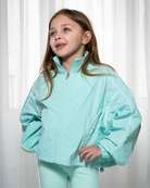 Young girl wearing a light blue jacket and pants against a white curtain background