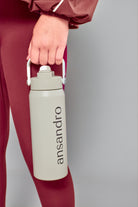 A person in burgundy activewear holds a light gray insulated water bottle with a white handle and the brand name 'ansandro' printed vertically in black on a plain gray background.