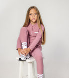 Young girl wearing a pink tracksuit sitting on a white stool against a light gray background